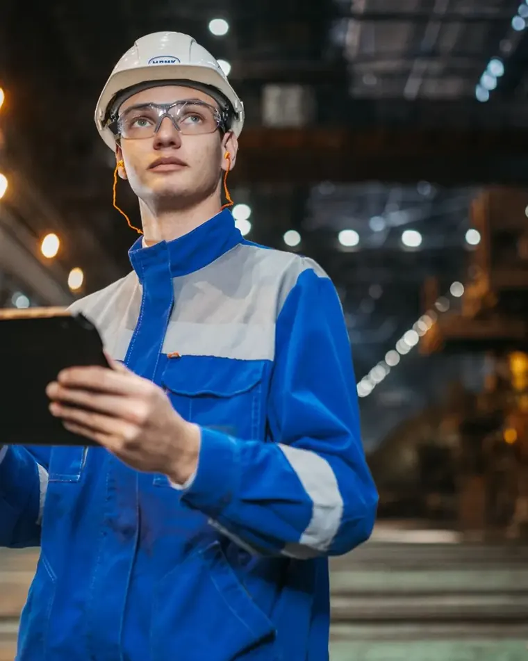 Engineer in a blue work jacket with reflective tape checking a tablet on the plant floor